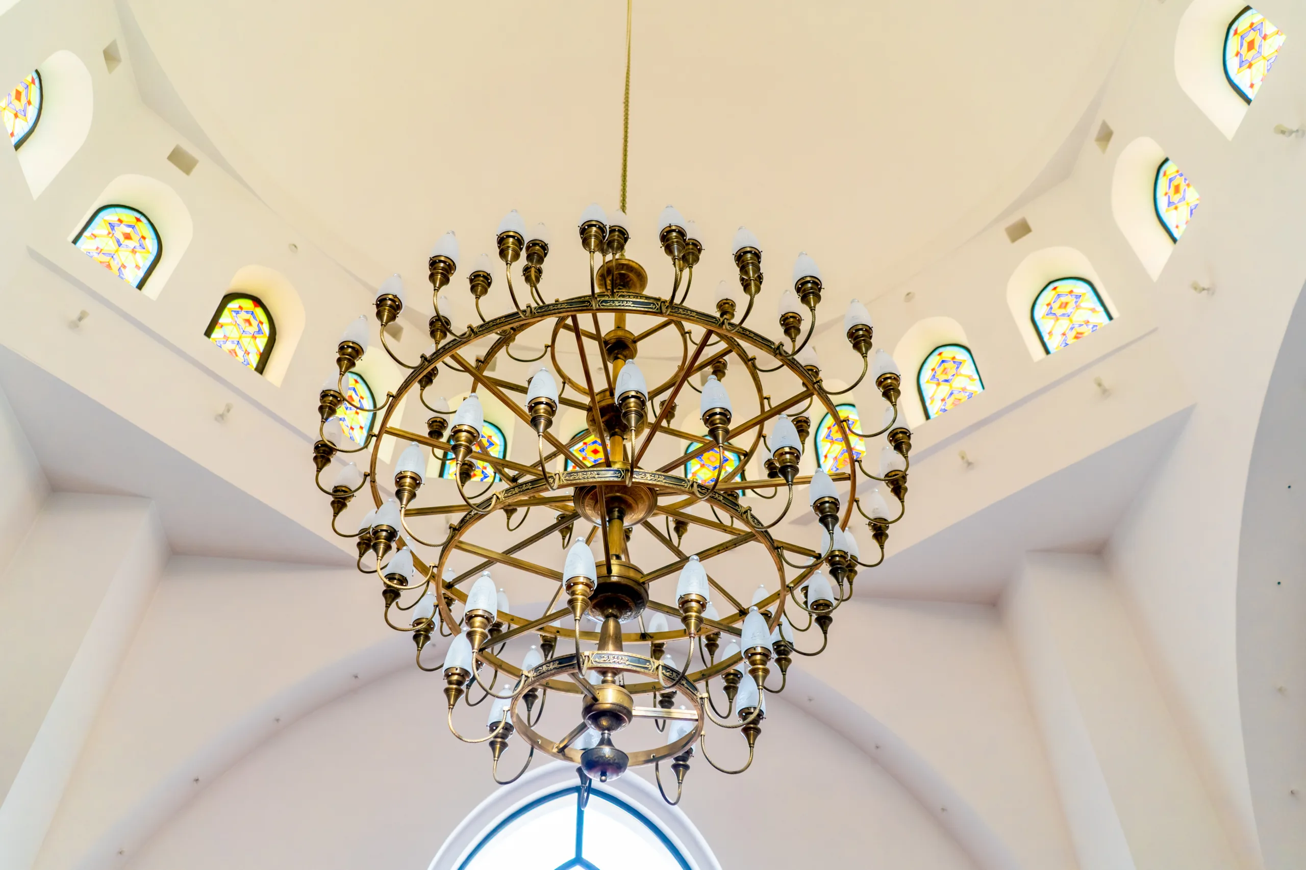 A grand, gold chandelier with multiple tiers and white bulb lights hanging from the ceiling of a hall with arched windows featuring colorful stained glass.